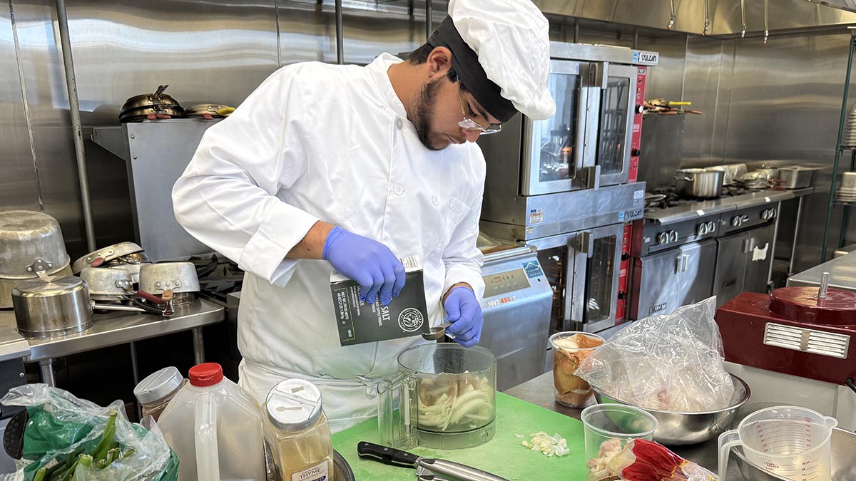 Culinary Student at prep station pouring salt into food processor bowl 